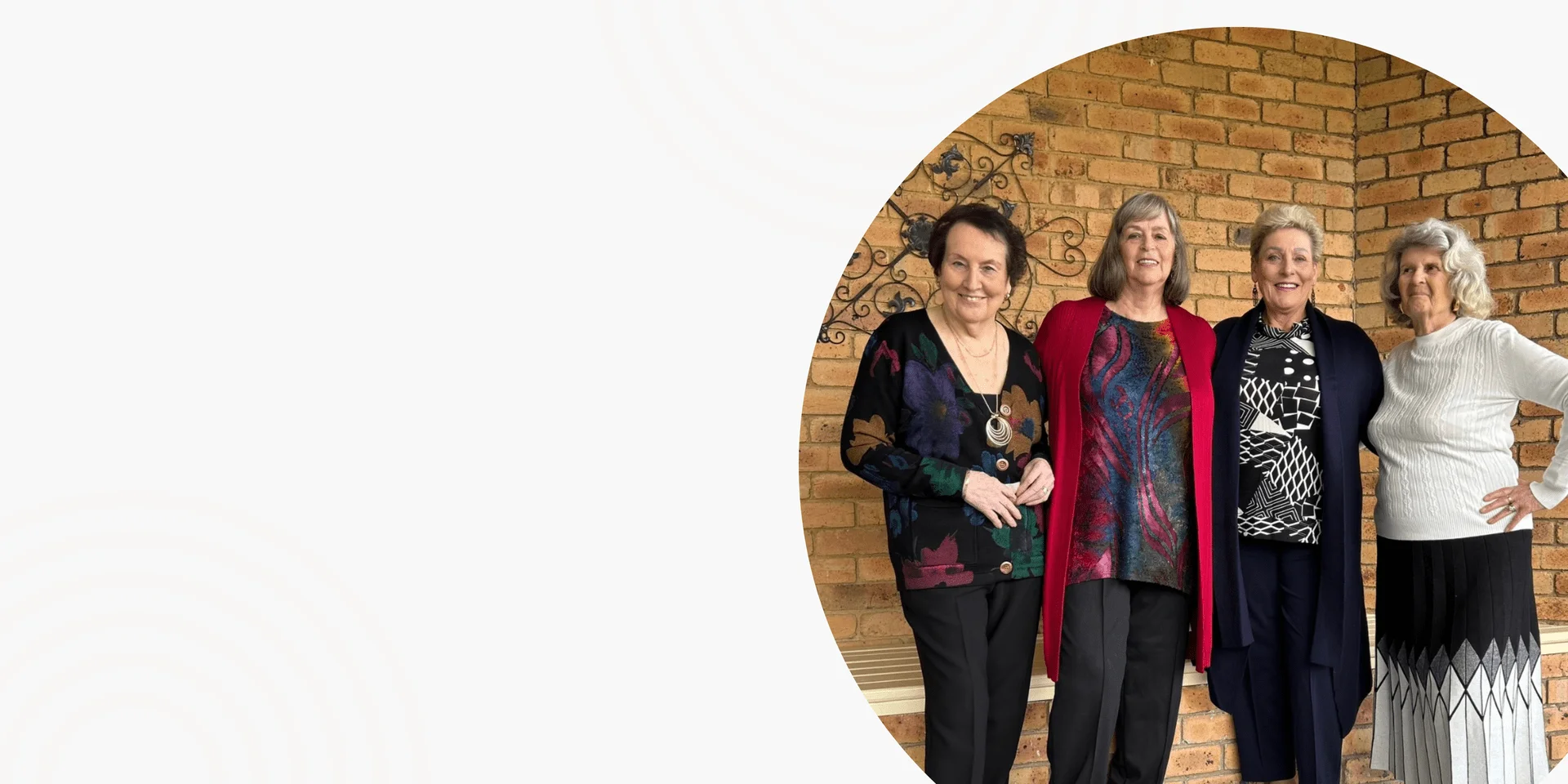 Four mature women standing in front of a red brick archway in black, red, monochrome print and cream knit outfits